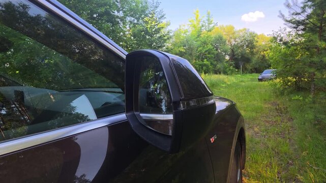 View of side mirror and front fender of a dark modern car parked on a grassy track, surrounded by lush green trees, under a clear blue sky with a distant vehicle, copy space