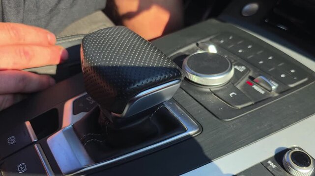 Interior view of modern car dashboard with black textured gear shifter and infotainment control panel illuminated by natural sunlight, driver hand visible.