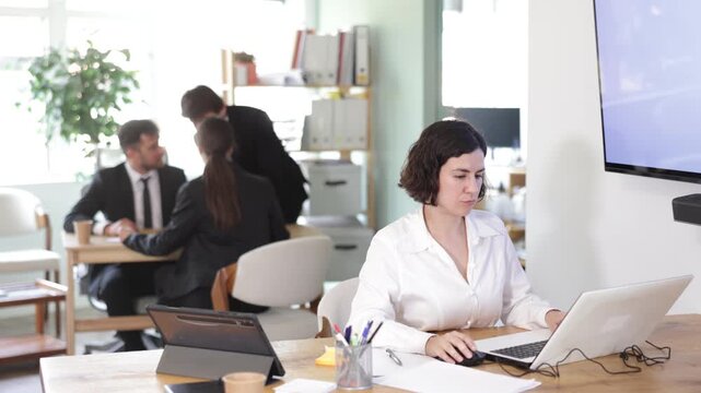 Adult woman sits at desk in office, focused on working with laptop during workday 