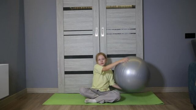 Young blonde girl sitting on yoga mat with fitness ball indoors, resting during home exercise routine, promoting healthy lifestyle for children, physical activity at residence