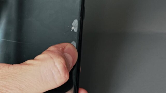 Close up view of male hand holding black smartphone with multiple impact cracks on protective screen film, displaying significant damage to mobile device display. Cracked screen protector