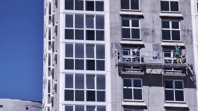 Two construction workers on suspended scaffold platform perform exterior finishing work on facade of tall modern residential building new construction site development clear blue sky