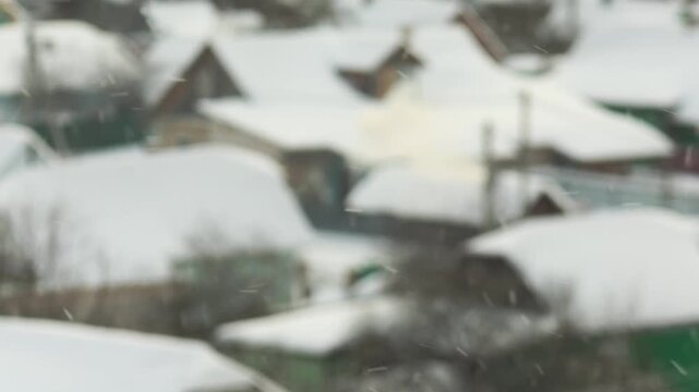 Winter scene of snow falling over village landscape, rooftops of houses covered in thick layer of white snow, creating peaceful wintertime atmosphere, slow motion