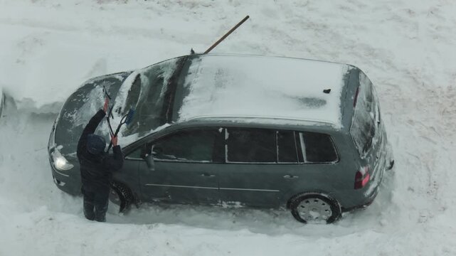 Person cleaning snow from windshield of car buried in deep snowdrift during heavy winter snowfall conditions, challenging winter morning scene, slow motion