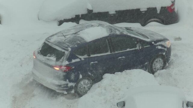 Dark blue hatchback car stuck in deep snow drift during severe winter snowstorm, wheels spinning furiously, driver attempting to free vehicle from harsh weather conditions on city street, slow motion