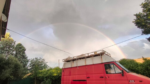 A vibrant double rainbow arching across a cloudy sky over a red van parked outdoors