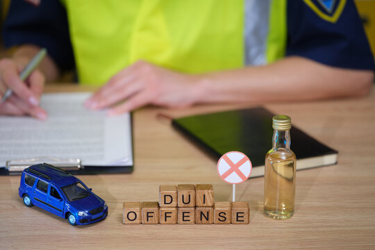 A police officer writes notes on a clipboard while a toy van and a small bottle sits beside wooden blocks spelling out DUI offense on a desk, emphasizing the serious nature of driving under influence.