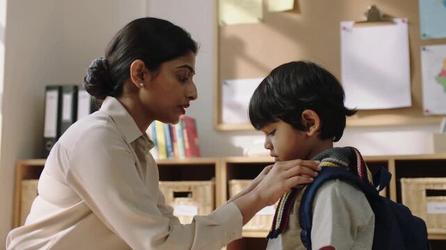 Approaching left woman in top adjusting boy's sweater and backpack straps in classroom checking fit