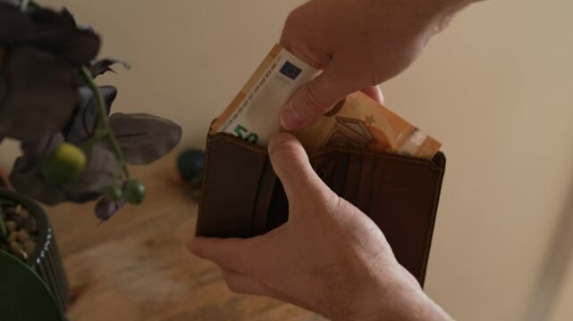Close up of male hands stuffing a stack of 50 euro banknotes into a brown leather wallet indoors. Useful for financial, banking, and wealth concepts.