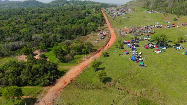 Rally car races and drifts along the red gravel road during the Nueva Alborada stage in Itapua, Paraguay, South America, passing spectators and campsites, drone forward flyover.