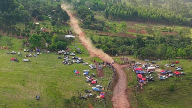Rally car speeds along a dirt road between spectators, parked vehicles and a temporary campsite in Nueva Alborada, Itapua, Paraguay, drone pullback.