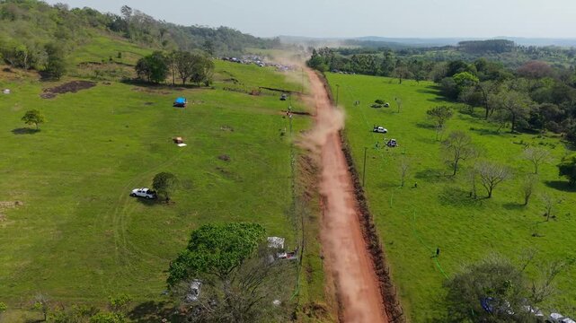 Rally car speeds along a long dirt special stage through green pasture and farmland in Nueva Alborada, Itapua, Paraguay, South America, Drone Forward Flyover.