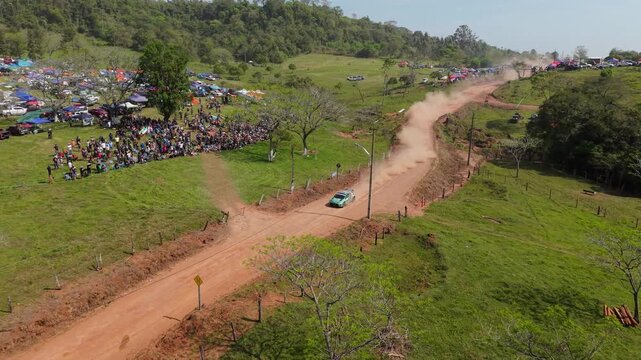 Rally cars speed along a winding dirt road, raising dust plumes beside spectators and campsites in Nueva Alborada, Itapua, Paraguay, South America, drone lateral tracking shot.