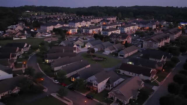 Dense mixed housing spreads across curved streets in Cornerstone, Pennsylvania, apartment buildings and single family homes glow warmly at dusk beneath dark wooded hills. Aerial drone orbit shot