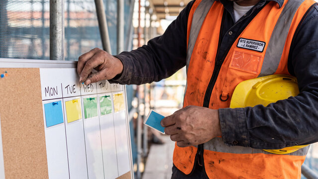 Construction project planning and schedule management. Site supervisor in safety vest organizing weekly task cards on board at building site for team workflow, scheduling and productivity