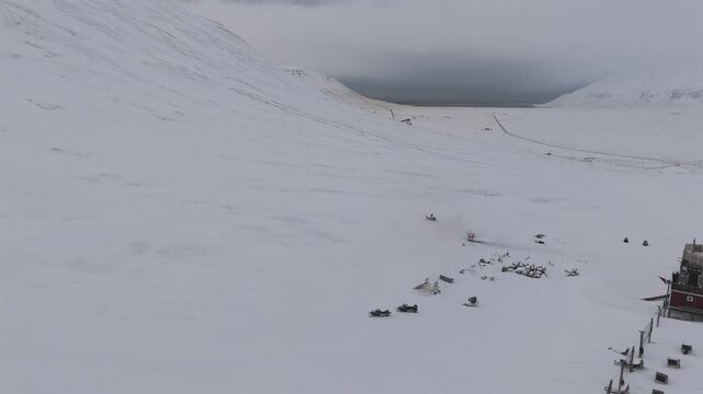 A snowmobile approaches a dog sled base with rows of tethering posts and parked sleds in an open Svalbard valley beneath low hanging clouds, aerial view.