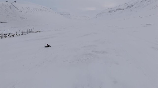 A snowmobile departs a dog sled base with rows of empty tethering posts in a wide, open snowy Arctic valley flanked by steep mountains, push forward aerial