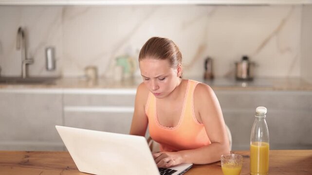 Thoughtful young girl looking confused at laptop computer near kitchen counter 