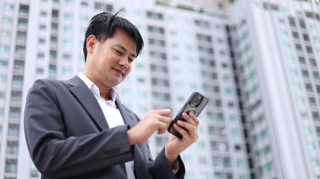 Asian businessman in suit using smartphone outdoors near city building. Low angle view of happy male executive texting on touch screen mobile phone, smiling during digital connection