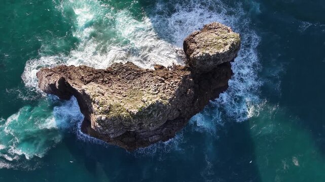 Aerial footage captures a dramatic rock sea stack off Oahu, Hawaii, as turquoise ocean waves churn and foam around rugged cliffs in bright daylight from a cinematic bird&rsquo;s eye view.