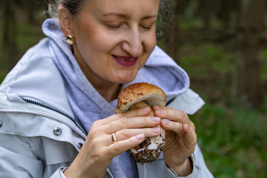 Portrait of mature woman smiling while smelling freshly picked porcini mushroom in forest. Concept of joyful sensory connection with nature during wild mushroom foraging in pine woodland setting.