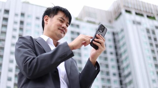 Asian businessman in suit using smartphone outdoors near city building. Low angle view of happy male executive texting on touch screen mobile phone, smiling during digital connection