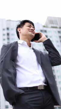 Asian businessman in suit talking on smartphone outdoors. Low angle view optimized for mobile display, social stories, business connection, and corporate mobile advertising