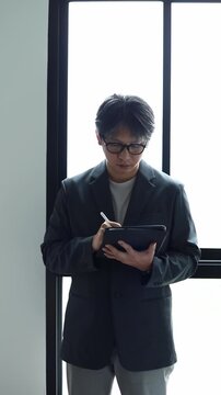 Asian businessman in suit writing on tablet with stylus pen indoors. Modern framing optimized for mobile display, social stories, business planning, and corporate ads