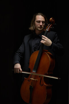 Vertical shot of young adult Caucasian musician playing cello instrument against dark studio backdrop. Classical performance imagery suited for music school advertising and concert promo