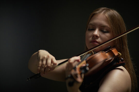 Young adult Caucasian woman playing violin during classical music performance. Visual asset representing focus and creativity for music school advertising and concert promotion
