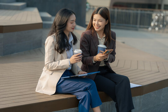 Two Asian businesswomen discussing work ideas over coffee with laptop and papers on wooden table.