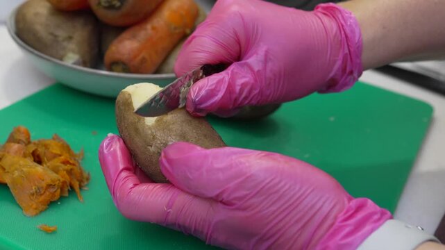 Pink gloved hands peeling raw Solanum tuberosum for healthy meal preparation. Macro shot focuses on knife blade removing skin from organic vegetable surface.