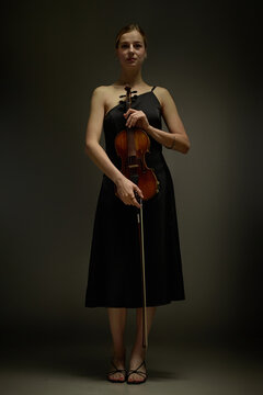 Vertical portrait of young adult Caucasian woman holding violin with bow, standing in studio. Suitable for classical music education, concert performance advertising