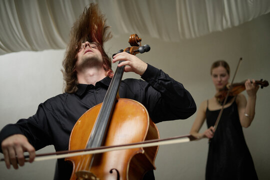 Caucasian young adult man playing cello with hair flying during passionate performance beside Caucasian young woman playing violin. Visual concept for music festival advertising