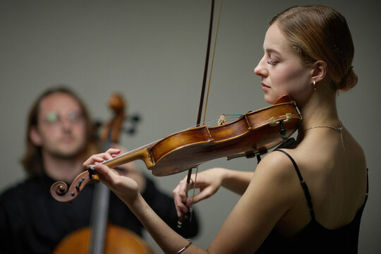 Young adult Caucasian woman playing violin during classical music rehearsal with cello player in background. Perfect for arts education, concert promotion and orchestra advertising