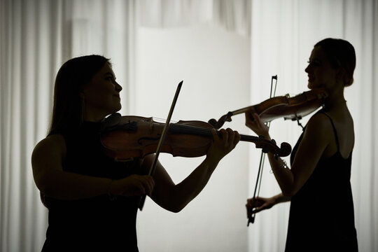 Two Caucasian young adult female musicians playing violins during classical concert rehearsal. Useful for music academy promotion, orchestra marketing, or arts festival advertising