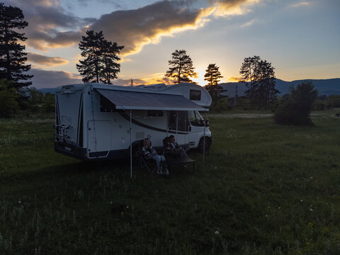 Couple enjoying the outdoor lifestyle, sitting under an awning next to their motorhome in a grassy field, watching the beautiful sunset behind distant pine trees while camping