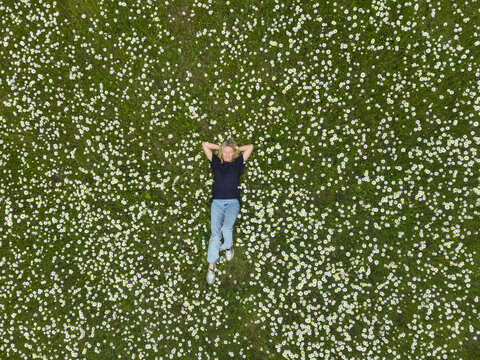 Woman lying on her back, hands behind her head, resting peacefully on a vibrant green field filled with numerous white daisies, enjoying nature during springtime