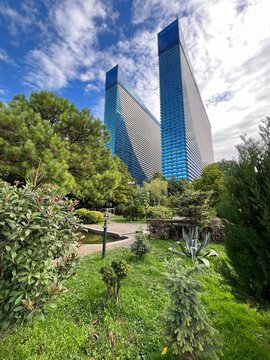 Low-angle view of Orbi City twin towers skyscrapers rising above a lush green park with trees, plants, and lawn under a blue cloudy sky in Batumi, Georgia.