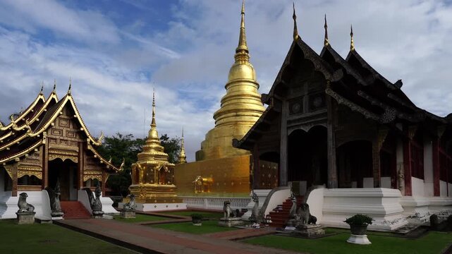 A wide-angle panoramic view capturing the majestic golden chedi spire alongside the intricately crafted wooden Lanna assembly chapels at Wat Phra Singh Worama in Chiang Mai, Thailand