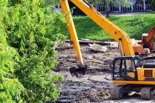 Two yellow tracked excavators operate on a muddy construction site, digging into dark, loose soil beside a paved walkway