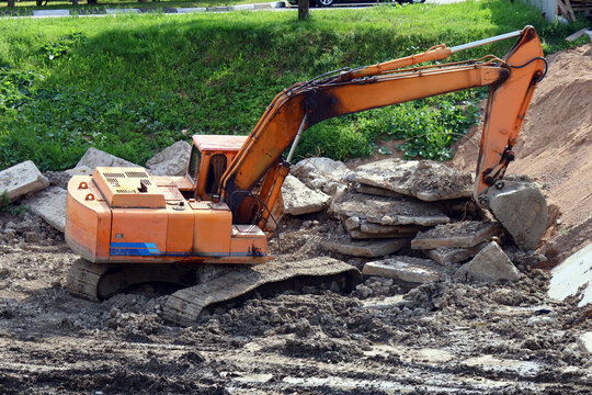 Orange tracked excavator works at a demolition site surrounded by broken concrete slabs and loose soil