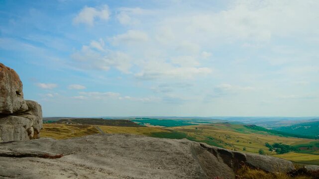 Expansive moorland panorama with gritstone outcrop and winding country road, slide shot. Broad cloud formations above rolling farmland and wooded hills. Rocky hillside viewpoint with cultivated fields
