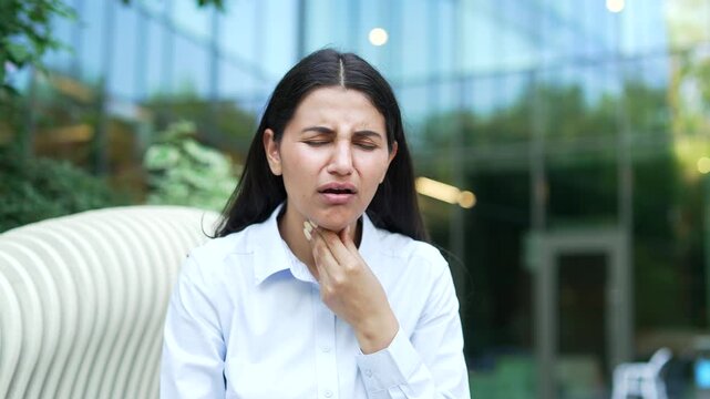 A businesswoman holding her neck in pain while suffering from a severe sore throat, sitting on a bench near office building outdoors. A sick female employee feeling tonsillitis agony outside. Close up