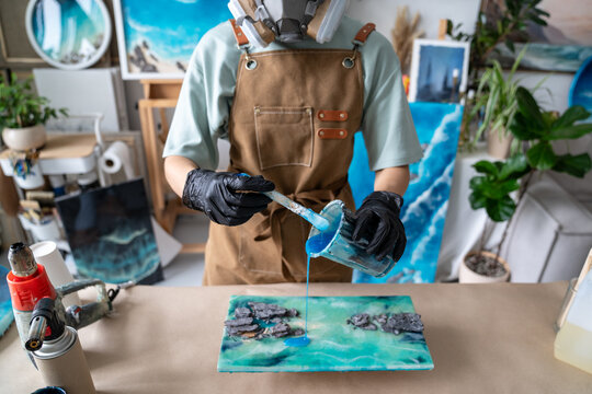 Woman artist wearing respirator and gloves mixes blue epoxy resin in cup at home loft worktable, preparing material for original handmade paintings produced and sold through creative small business.