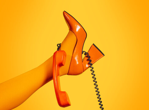 Leg in orange heel with retro phone receiver on orange background