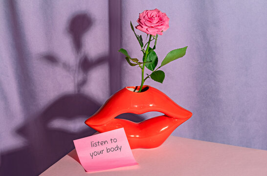 A red lip vase holds a rose near a listen to your body note