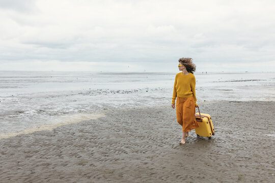 A girl walks barefoot on the beach pulling a yellow suitcase