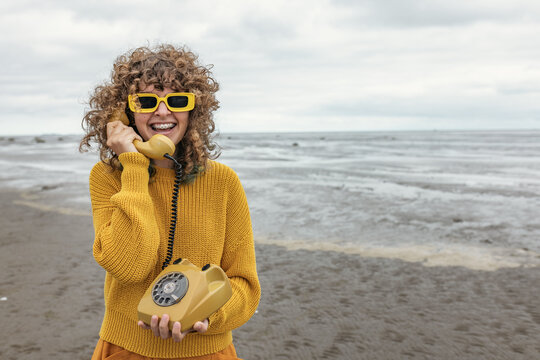 Smiling girl in yellow sweater talks on retro telephone at the beach