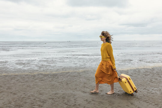 Woman pulls a yellow suitcase while walking barefoot on the beach